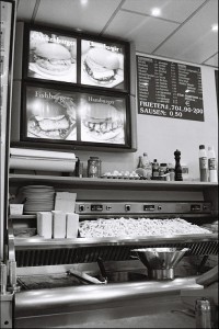 A black and white photo of a fast food restaurant counter in Ostende displays a menu board with items like Chickenburger, Fishburger, and Hamburger. Below the menu, there are plates, condiment bottles, and a tray of freshly cooked French fries, capturing an errance in culinary simplicity.